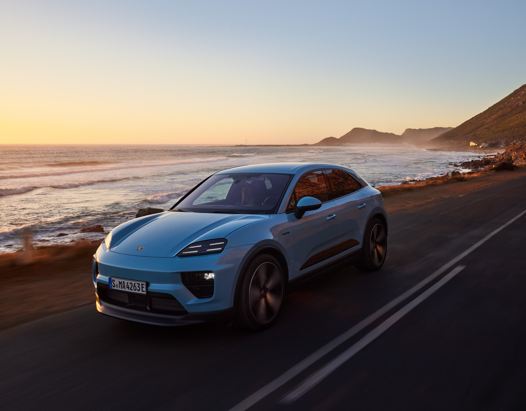2025 Macan Electric in Frozen Blue Metallic driving on oceanside road with waves crashing in the background