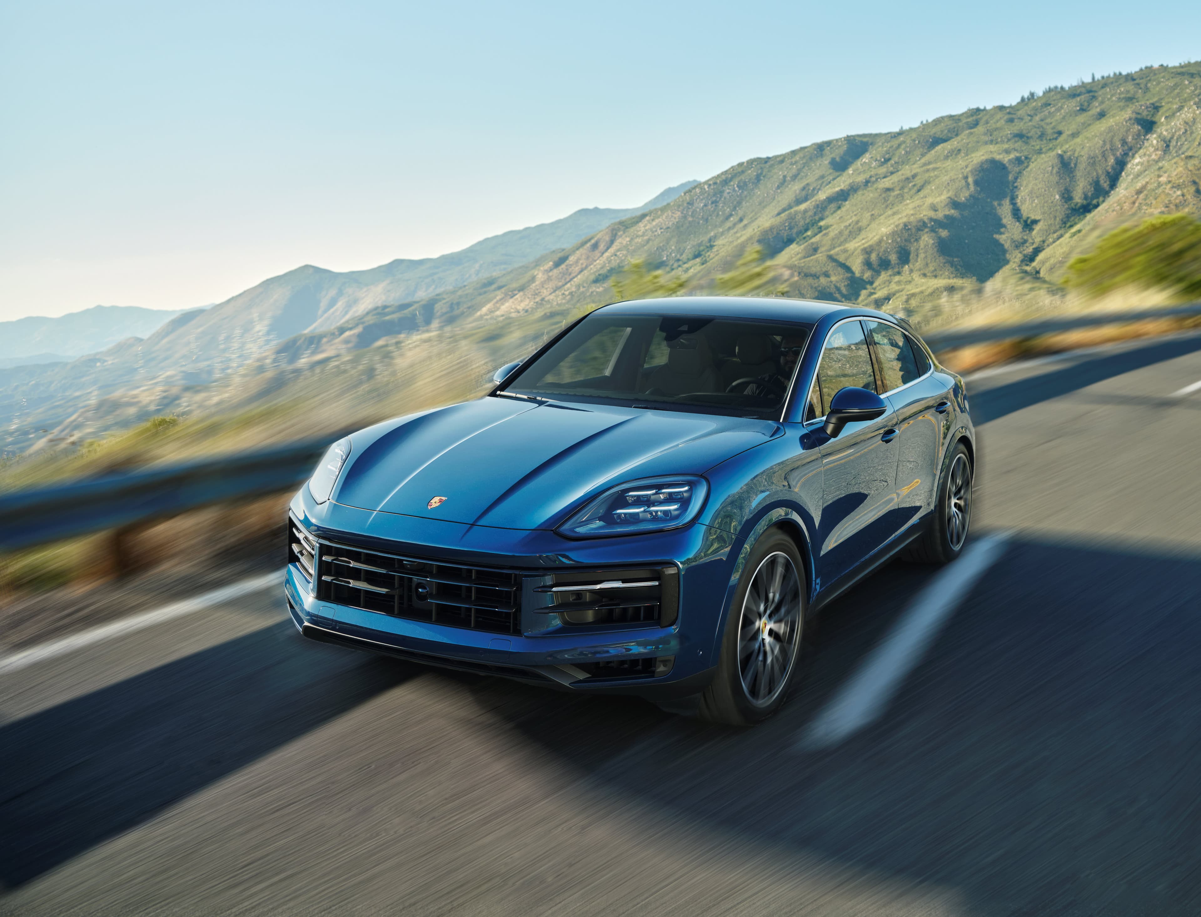 A blue 2023 Cayenne S Coupé drives on a scenic mountain road, with blurred motion and green hills in the background under a clear blue sky.